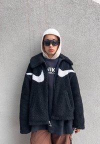 Μαύρο φλις μπουφάν με λευκά λογότυπα Nike, κλείσιμο με φερμουάρ και σχέδιο με γιακά, το οποίο είναι επενδυμένο πάνω από μια μαύρη μπλούζα και καφέ παντελόνι.