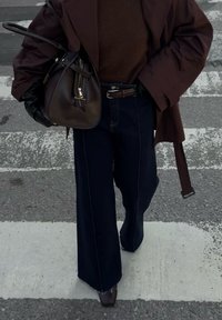 Brun oversized yttertøy, brun strikket topp, mørk denim med vid ben, svarte lærhansker, brun håndveske med glidelås, står på et fotgjengerfelt.