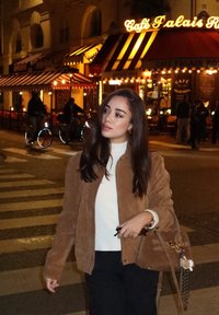 Brun mockajacka över en vit topp, matchad med svarta byxor. Accessoarer inkluderar en ljusbrun handväska med gulddetaljer och ett berlock.