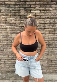 Musta lyhyt paita, vaaleansiniset korkeavyötäröiset farkkushortsit, joissa on rispaantuneet reunat. Pitää käsissään mustavalkoisia aurinkolaseja, seisoo kiviseinää vasten.