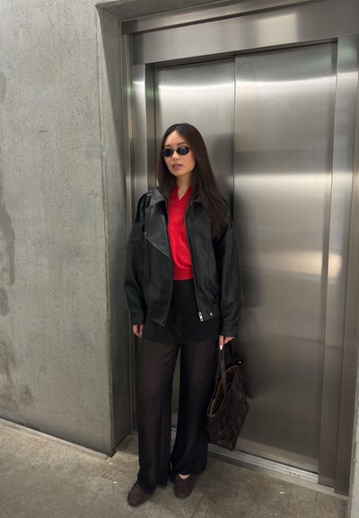 Mujer con chaqueta de cuero negra, top rojo, pantalones anchos negros y gafas de sol, de pie sosteniendo una bolsa marrón cerca de una puerta de ascensor cerrada.