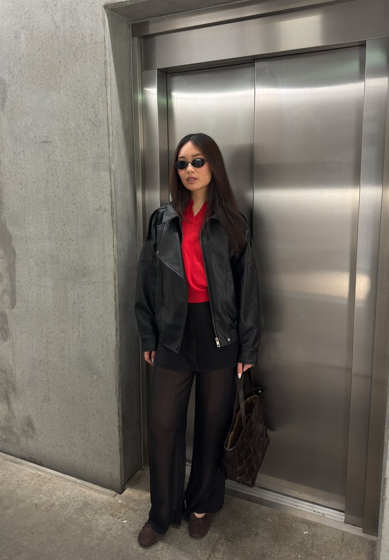Woman wearing black leather jacket, red top, black wide pants, and sunglasses stands holding a brown bag near a closed elevator door.