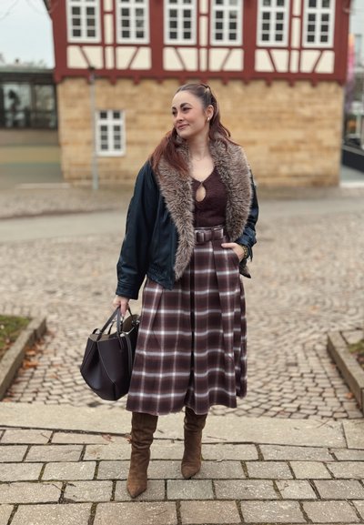 Falda midi de cuadros marrones con cinturón en la cintura, combinada con una chaqueta bomber negra con cuello de piel. Botas marrones hasta la rodilla y un bolso negro.