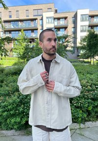 Camicia beige chiaro a maniche lunghe con chiusura a bottoni, tasca frontale e branding sottile. Indossata sopra una t-shirt scura, abbinata a pantaloni corti chiari.