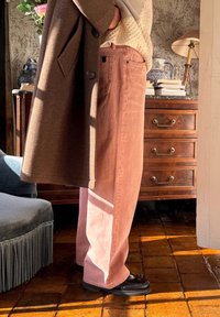Svetlo roza široke hlače iz mehkega denima, v kombinaciji z teksturiranim rjavim plaščem. Črne čevlje dopolnjujejo oblačila. Ozadje prikazuje leseno pohištvo.