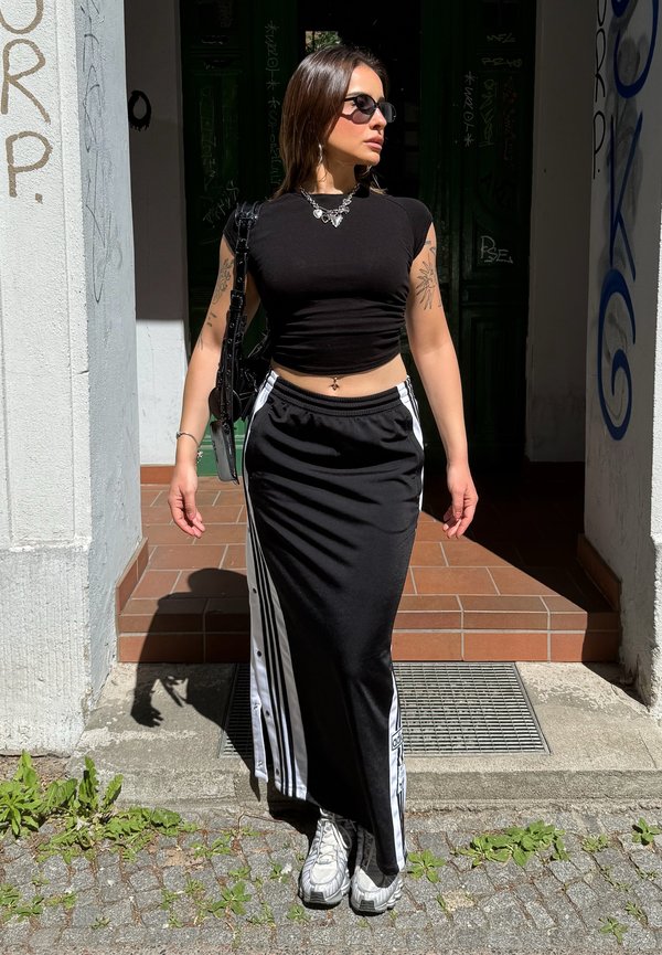 Femme portant un crop top noir, une longue jupe noire et blanche, des baskets blanches, des lunettes de soleil et un collier, debout au soleil près de l'entrée d'un bâtiment.