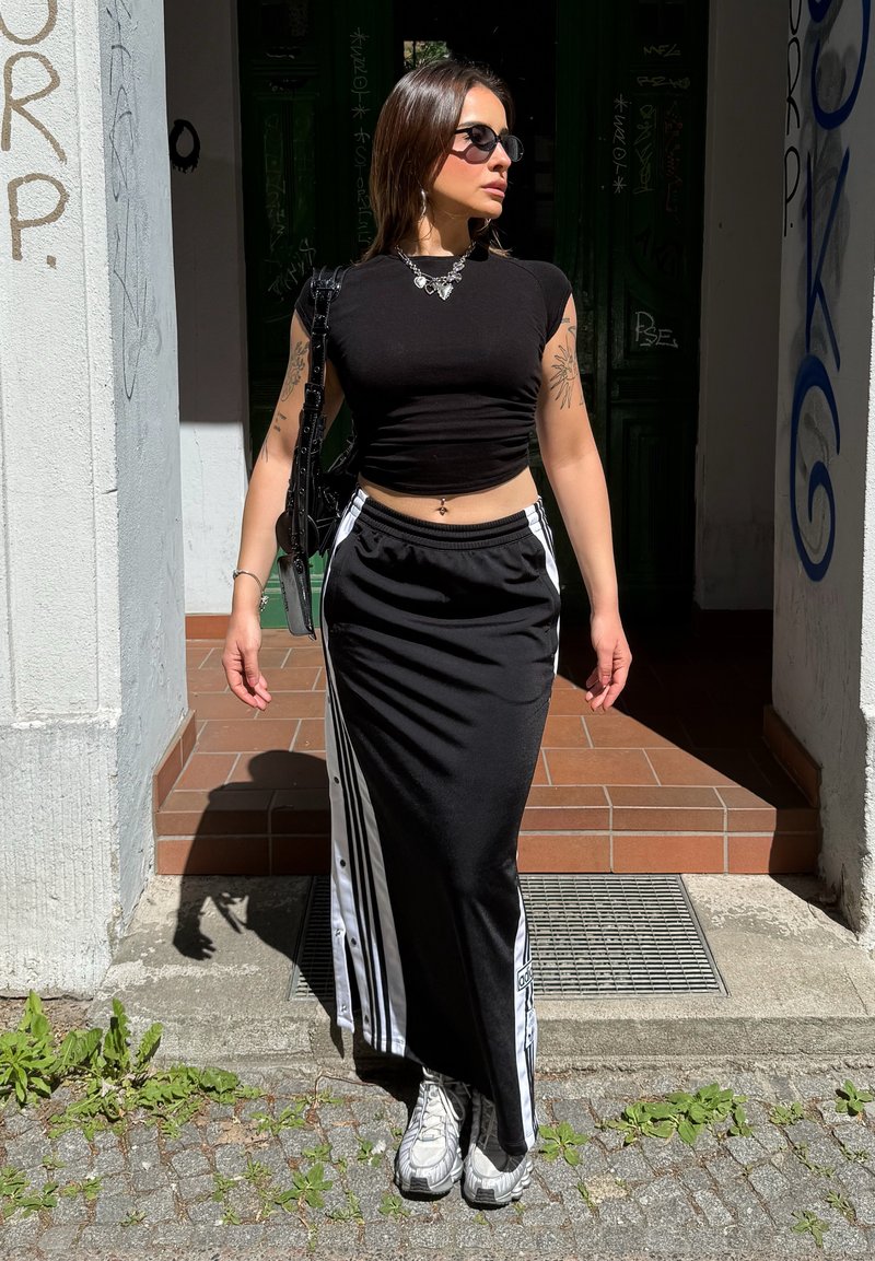 Femme portant un crop top noir, une longue jupe noire et blanche, des baskets blanches, des lunettes de soleil et un collier, debout au soleil près de l'entrée d'un bâtiment.