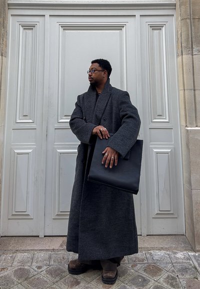 Hombre con un largo abrigo gris oscuro y gafas, sosteniendo una gran bolsa negra, de pie frente a una puerta blanca panelada en una calle adoquinada.