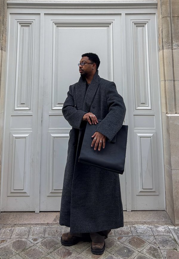 Homme portant un long manteau gris foncé et des lunettes, tenant un grand sac noir, debout contre une porte blanche à panneaux sur une rue pavée.