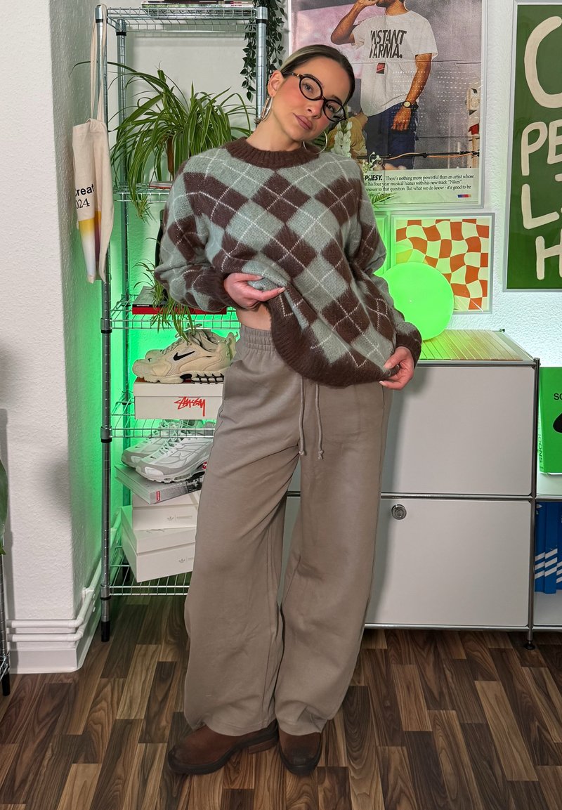 Knitted sweater in teal and brown argyle pattern, paired with beige wide-leg pants. Accessories include large glasses and brown footwear.