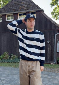Suéter de punto a rayas en azul marino y blanco con cuello redondo, combinado con pantalones beige claros. El suéter tiene un detalle del logo de Lacoste en el pecho.