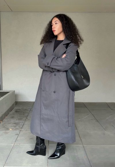 Abrigo largo gris tipo gabardina con botones dobles, combinado con botas negras de tacón alto y un bolso de hombro negro. Acabado de tela lisa.