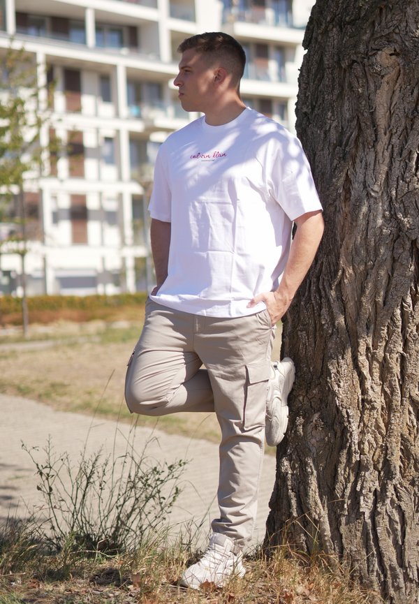 Joven vestido con camiseta blanca y pantalones cargo beige se apoya en un árbol con una pierna doblada en un parque urbano al aire libre.