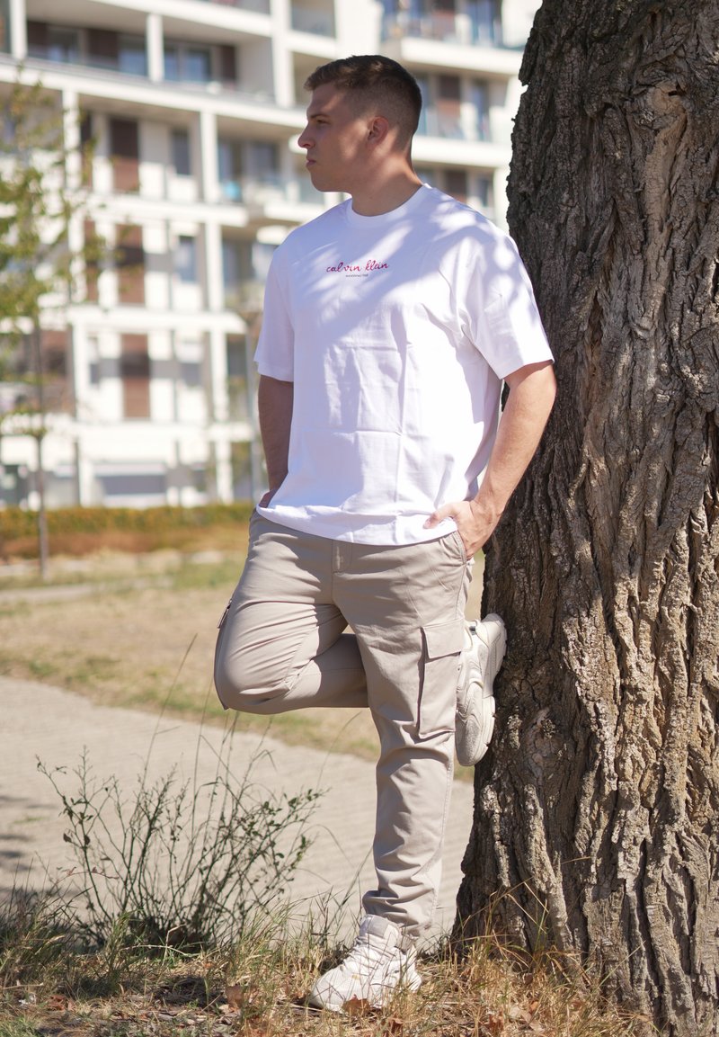 Jeune homme en t-shirt blanc et pantalon cargo beige appuyé contre un arbre, une jambe pliée, dans un parc urbain en plein air.