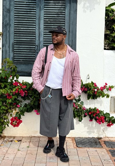 Camisa de botones a rayas rosas sobre una camiseta sin mangas blanca, pantalones cortos de rayas grises, zapatillas negras, detalle de cadena, gafas de sol, con flores de fondo.