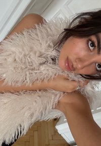 Étole en fourrure beige clair, duveteuse et à poils longs, drapée sur une épaule, avec une surface texturée et un gros plan sur une main la tenant.