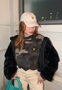 Νεαρή γυναίκα που φοράει μπεζ καπέλο και καμουφλαρισμένο πουλόβερ κάτω από μαύρο γούνινο μπουφάν, κρατώντας τυρκουάζ τσάντα, στέκεται μέσα σε εσωτερικό χώρο κοντά σε παράθυρο.