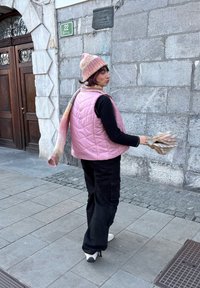 Veste matelassée rose portée sur une chemise noire à manches longues, associée à un pantalon cargo noir et un bonnet et une écharpe tricotés multicolores.