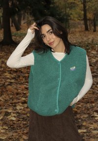 Turkusowa polarowa kamizelka z okrągłym dekoltem, teksturowym materiałem i kontrastującymi jasnoniebieskimi guzikami. Kamizelka jest noszona na białej koszuli z długim rękawem.