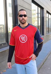 Sarkans un tumši zils sporta krekls ar īsām piedurknēm, dzelteniem akcentiem un apļveida "90" logo priekšpusē. Nēsāts virs garā melnā krekla.