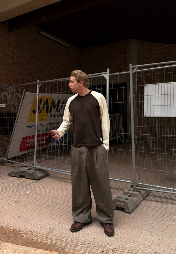 Man draagt losse broek en een shirt met lange mouwen, staat met een telefoon vastgehouden bij een metalen bouwschutting in een stedelijke omgeving.