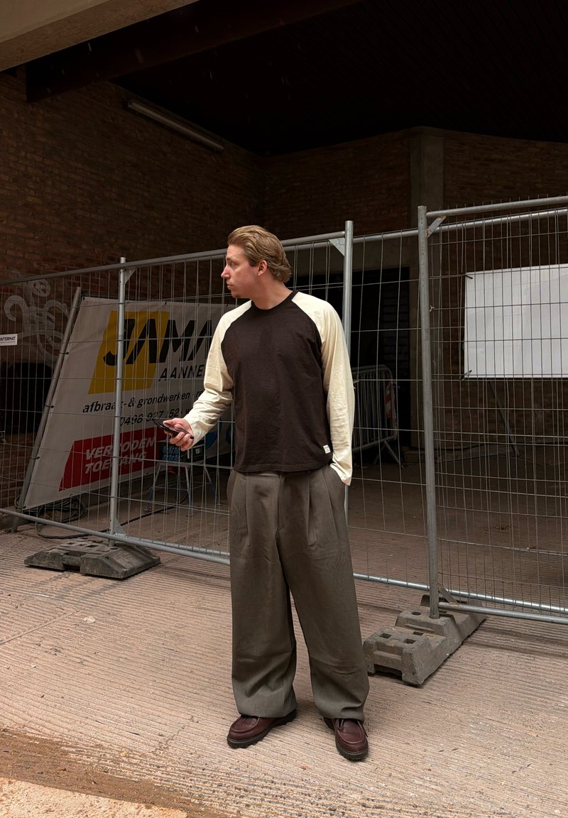 Muž oblečený v voľných nohaviciach a tričku s dlhým rukávom stojí a drží telefón pri kovovom plotovom konštrukcii v mestskom prostredí.