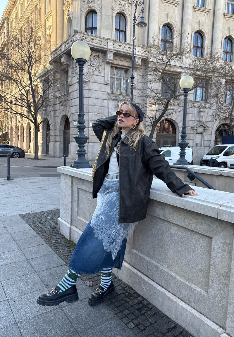 Femme blonde portant des lunettes de soleil, une veste noire, une jupe en dentelle sur du denim, des chaussettes rayées et des mocassins, appuyée sur une rampe en pierre dans un cadre urbain avec un bâtiment historique.