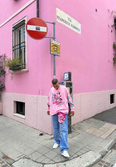 Camisa rosa de manga larga con diseño gráfico y detalles azules, combinada con jeans de mezclilla azul claro y deportivas blancas. Bolsa rosa y fondo de pared rosa.