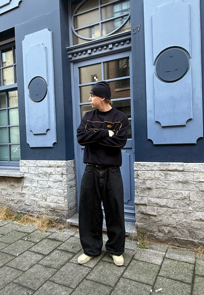 Sudadera negra con rayas amarillas, jeans negros holgados y zapatos beige, de pie frente a una fachada de edificio gris y azul.