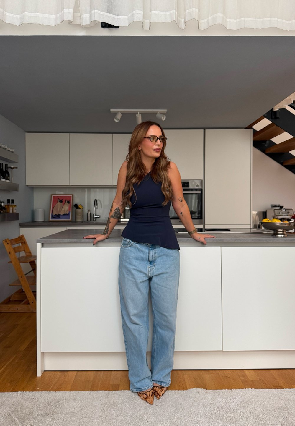 Persoon draagt een marineblauwe peplumtop en lichtblauwe jeans, staat bij een modern kookeiland met een grijs werkblad en minimalistische kasten.