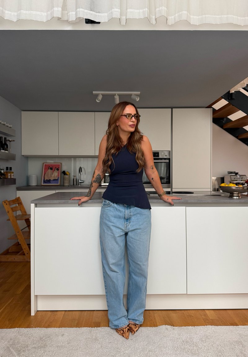 Personne portant un haut péplum bleu marine et un jean bleu clair se tient près d'un îlot de cuisine moderne avec un plan de travail gris et des armoires minimalistes.