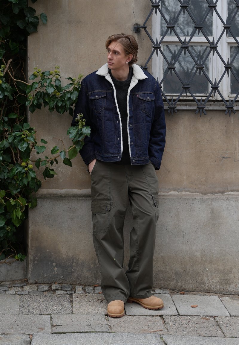 Jeune homme debout, les mains dans les poches, portant une veste en denim foncé avec col blanc, un pantalon cargo vert et des bottes beige, appuyé contre un mur de pierre.