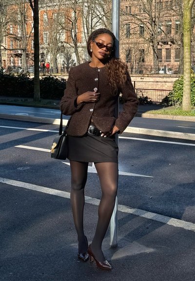 Mujer con gafas de sol oscuras, chaqueta marrón texturizada, minifalda negra y medias transparentes, de pie en una calle cerca de un poste a la luz del día.