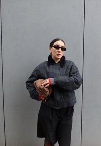 Une femme portant des lunettes de soleil foncées et une veste sombre tient un chapeau marron plié, debout contre un mur gris uni.