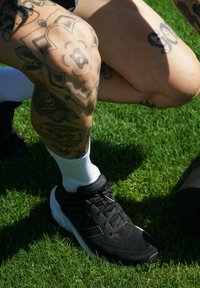 Zapatilla deportiva negra con parte superior texturizada y suela blanca, combinada con calcetines blancos de caña corta. Tatuajes visibles en la rodilla y piel expuesta sobre un fondo de hierba.