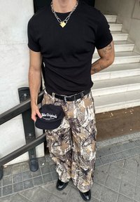 Camiseta preta justa, calças cargo em padrões de camuflagem com tons terrosos, pernas largas, cinto preto, segurando um boné preto com o logótipo "Adidas Originals".