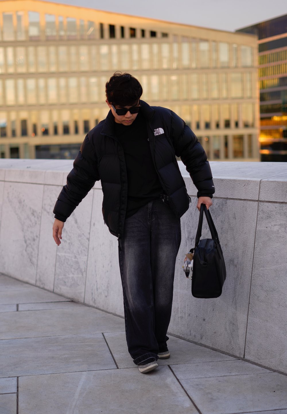 Μαύρο μπουφάν με λογότυπο, μαύρο T-shirt, φαρδιά μπλέ τζιν και μαύρα αθλητικά. Κρατάει μια μαύρη τσάντα και μια κάμερα. Υπαίθριο περιβάλλον.