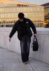 Μαύρο μπουφάν με λογότυπο, μαύρο T-shirt, φαρδιά μπλέ τζιν και μαύρα αθλητικά. Κρατάει μια μαύρη τσάντα και μια κάμερα. Υπαίθριο περιβάλλον.