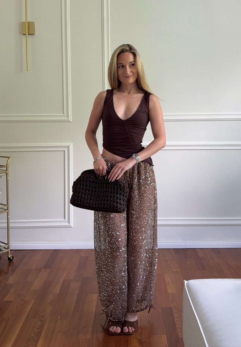 Femme debout à l'intérieur, portant un haut sans manches foncé, un pantalon large à sequins, tenant un sac à main tricoté, sur un sol en bois avec des murs clairs.