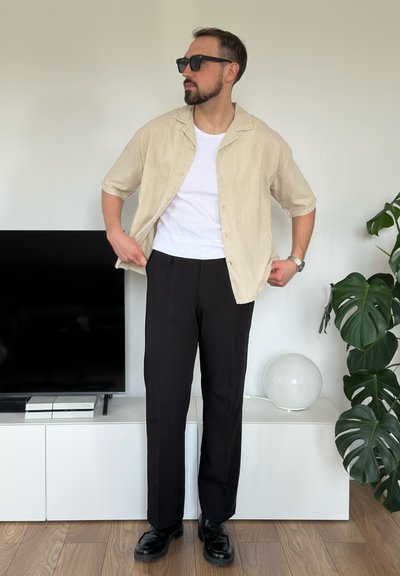Camisa de manga corta beige sobre una camiseta blanca, combinada con pantalones negros y zapatos negros; diseño moderno con textura suave.