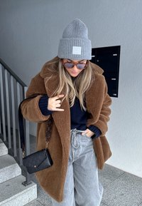 Brūns pūkains pārāk liels mētelis, tumši zils džemperis, pelēks beanie, saulesbrilles un gaiši pelēkas plaša piegriezuma bikses. Melna plecu soma ar logotipa detaļu.