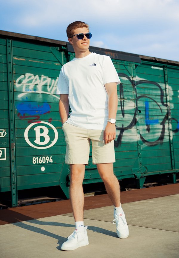 Maglietta bianca a maniche corte con un piccolo logo, pantaloni corti beige e sneakers bianche. Sullo sfondo si vede un container verde con graffiti.