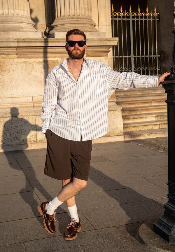 Bărbat purtând ochelari de soare, cămașă cu dungi, pantaloni scurți maro și pantofi, sprijinit de un stâlp de iluminat aproape de o clădire clasică din piatră la soare.