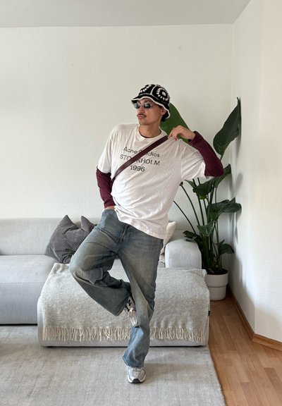 Hombre con sombrero de cubo, gafas de sol, camisas superpuestas, jeans y zapatillas posa en interiores cerca de un sofá y una planta alta, equilibrándose sobre una pierna.