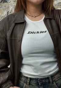 Ruda odinė striukė per baltą raukšlėtą tank top'ą su juodu tekstu "Dickies." Papuošalai apima sluoksniuotas auksines grandinaites.