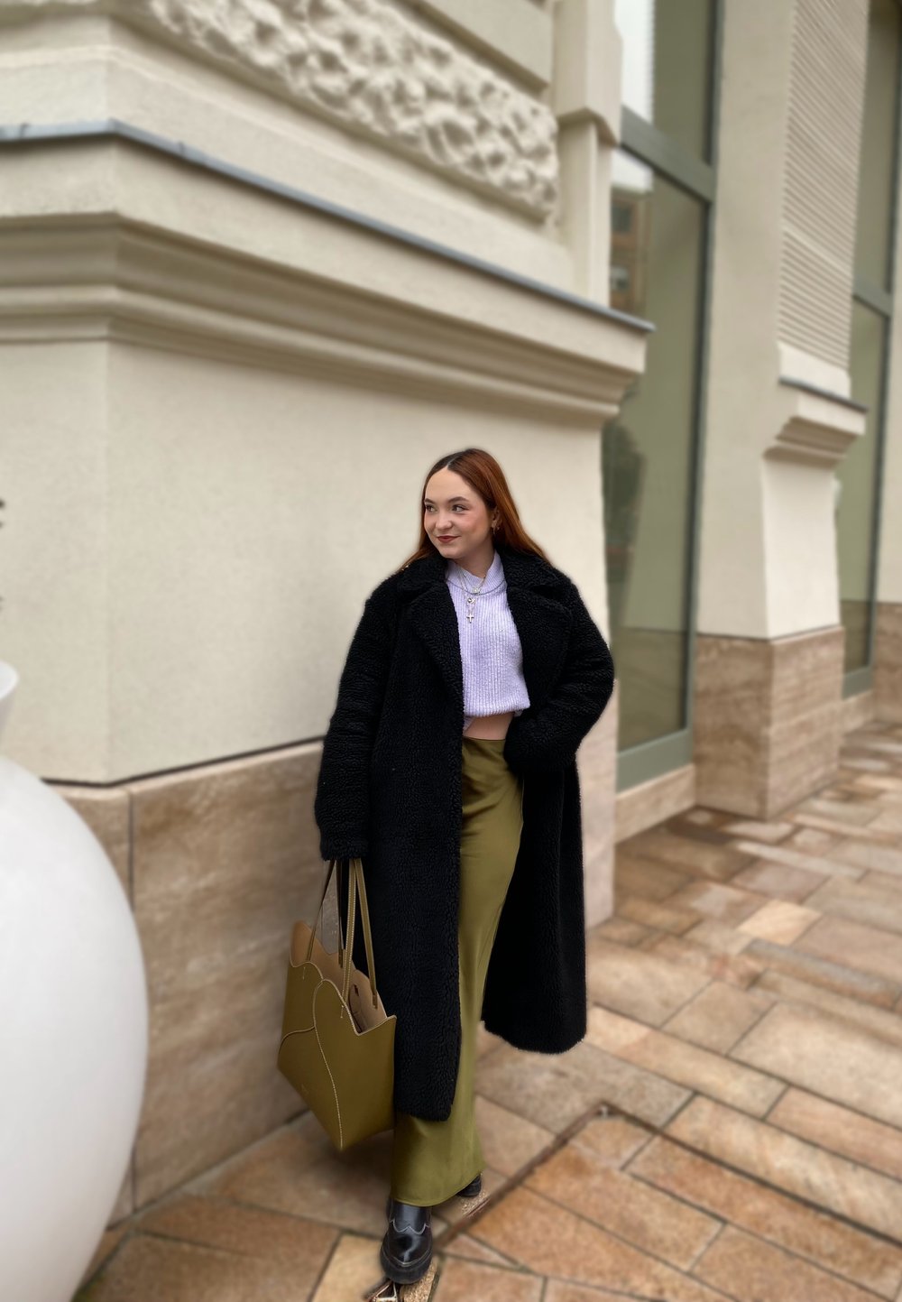 Čierny chundelatý kabát, svetlofialový skrátený sveter, olivovo zelená sukňa, čierna obuv a béžová nákupná taška, postavená pri textúrovanej stene.