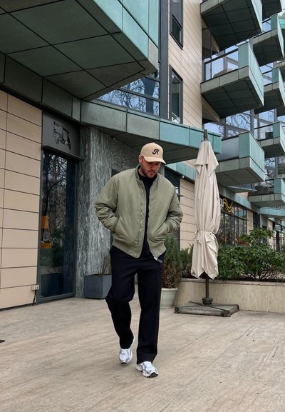Chaqueta bomber verde oliva clara, pantalones negros, zapatillas blancas y una gorra marrón. El fondo presenta arquitectura moderna y vegetación.