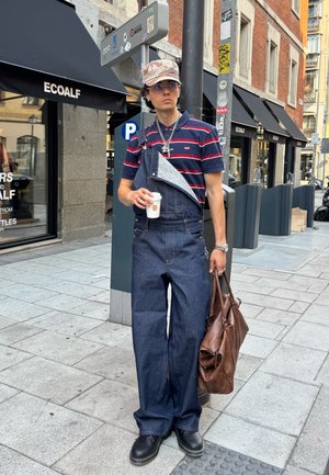 Pantalones de mezclilla con una tira caída, bolsillo gris, zapatos negros, camisa a rayas azul marino y roja, bolsa de cuero marrón, sosteniendo una taza de café.