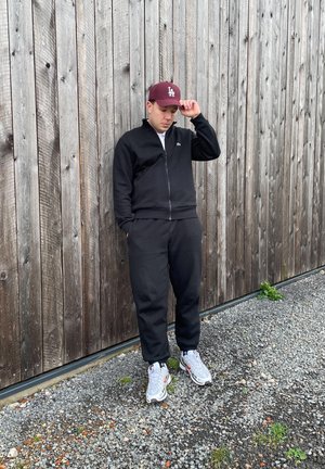 Chaqueta negra con cremallera y pantalones a juego, con zapatillas deportivas blancas y grises con estampado. Gorro burdeos con letras blancas, de pie frente a una pared de madera.