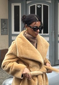 Μπεζ ψεύτικο γούνινο παλτό με φαρδύ γιακά και ζώνη στη μέση, φορεμένο πάνω από έναν καφέ ρόλο. Μαύρα γυαλιά ηλίου, χρυσά σκουλαρίκια και δαχτυλίδι προσθέτουν ένταση στην εμφάνιση.